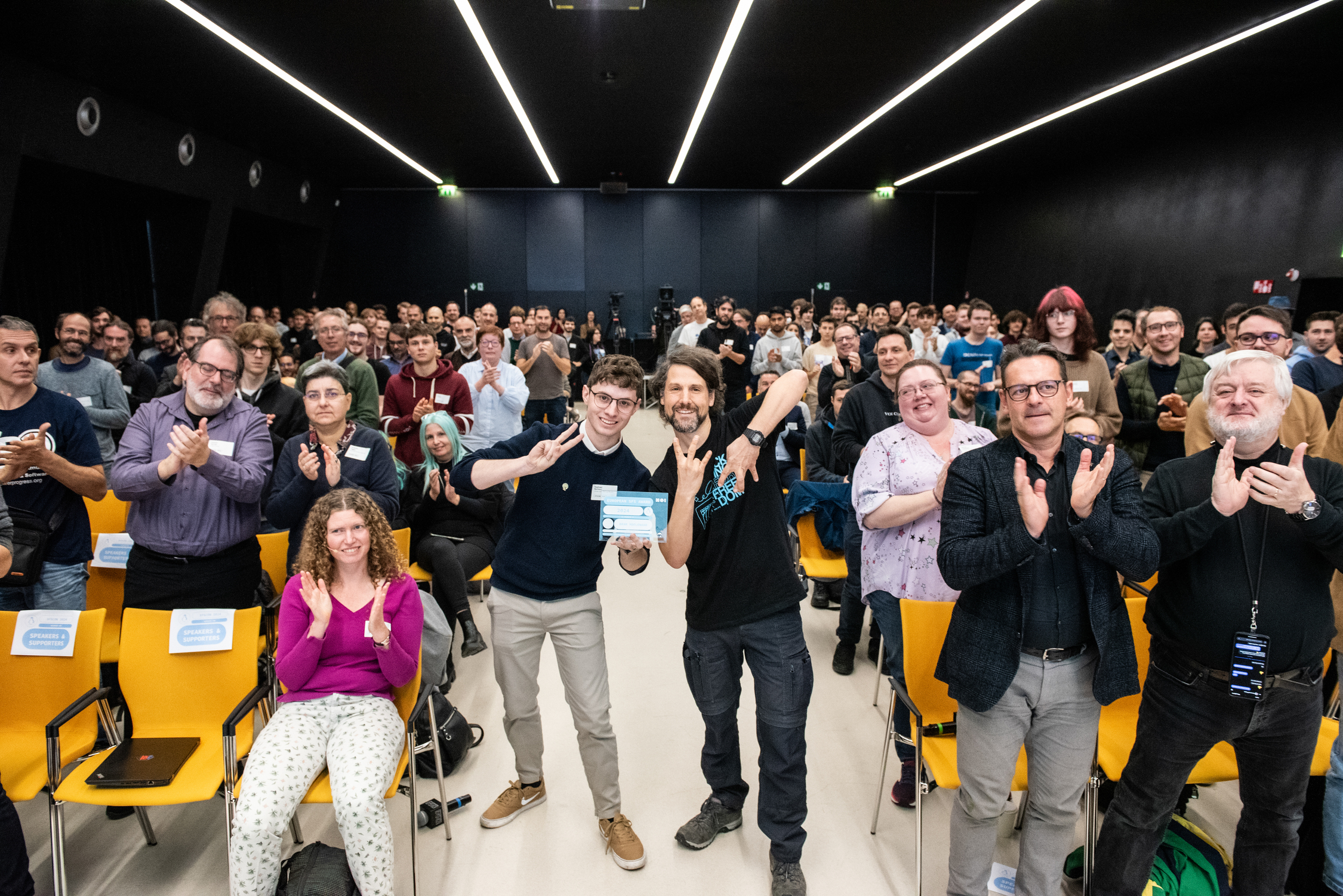 Matthias and Raphael at SFSCON 2024 asking all people in the audience
to stand up if are using Vim, or have used Vim in the past. Matthias
showing the VI M sign with his fingers - CC-BY-SA 4.0
NOI